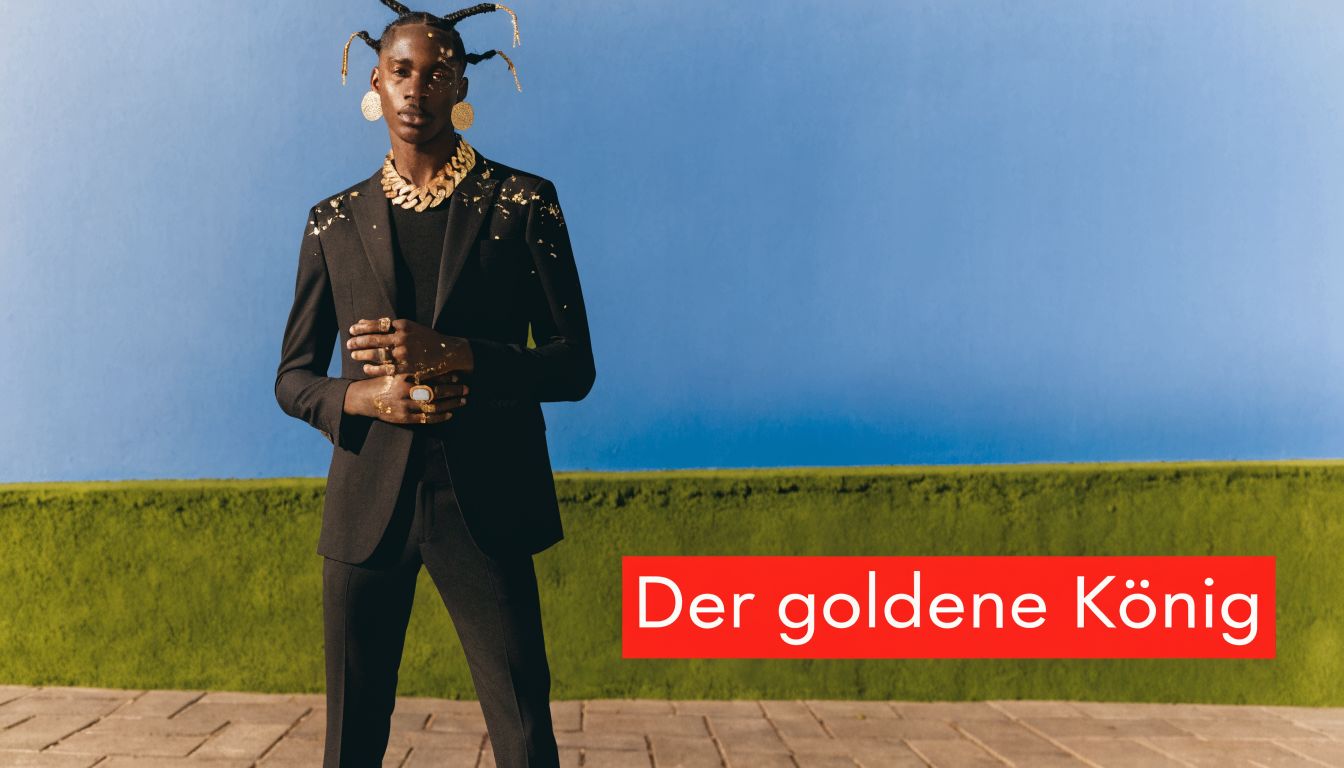 Ein stilvoller Mann mit Goldakzenten trägt einen eleganten schwarzen Anzug vor einer markanten blau-grünen Wand.